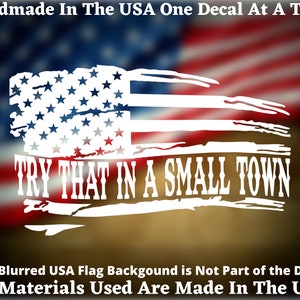 May include: White vinyl decal with a distressed American flag design and the text "TRY THAT IN A SMALL TOWN". The decal is on a blurred background of an American flag.