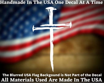 May include: White vinyl decal of a cross with nails, against a blurred American flag background. The text "Handmade In The USA One Decal At A Time" and "All Materials Used Are Made In The USA" are also included.