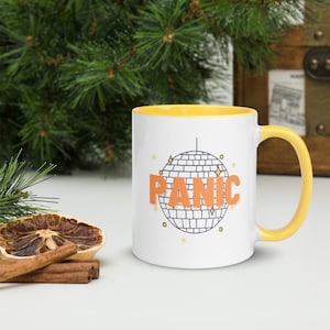 May include: White ceramic mug with a yellow rim and handle. The mug features a black and white illustration of a disco ball with the word "PANIC" in orange letters.