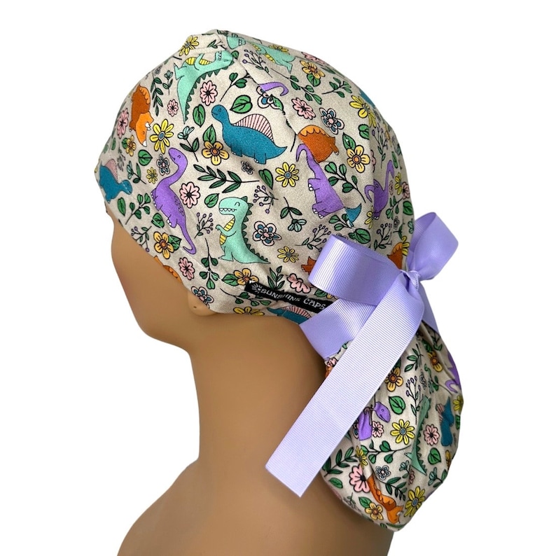 May include: A beige scrub cap with a colorful dinosaur and floral print. The cap has a purple ribbon tied at the back.
