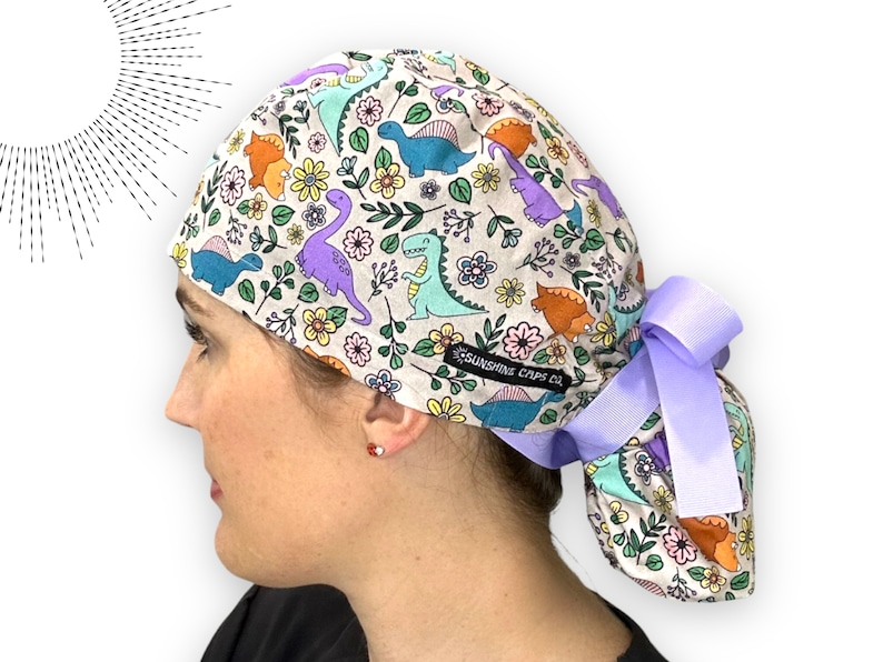 May include: A white scrub cap with a colorful dinosaur and floral print. The cap has a purple tie and the text "SUNSHINE CAPS CO." is printed on the side.