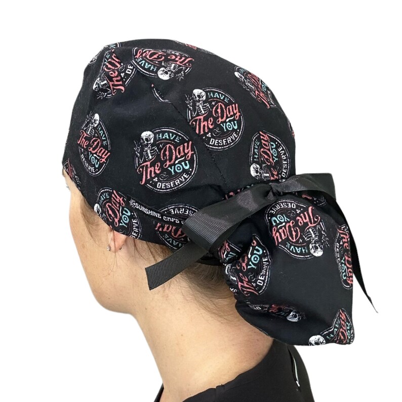 Puede incluir: Gorro quir&uacute;rgico negro con un dise&ntilde;o repetido que presenta un esqueleto, las palabras "Have The Day You Deserve" y un lazo negro. El gorro se lleva en la cabeza.