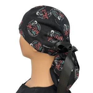 Puede incluir: Gorro quir&uacute;rgico negro con un patr&oacute;n repetido de gr&aacute;ficos de esqueletos y el texto "The Day You Deserve" en un dise&ntilde;o circular. El gorro tiene un lazo negro en la parte posterior.
