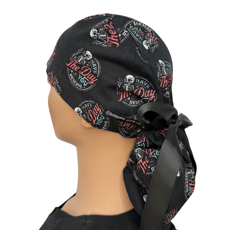 Puede incluir: Gorro quir&uacute;rgico negro con un patr&oacute;n repetido de gr&aacute;ficos de esqueletos y el texto "The Day You Deserve" en un dise&ntilde;o circular. El gorro tiene un lazo negro en la parte posterior.