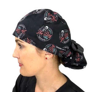 Puede incluir: Gorro quir&uacute;rgico negro con un patr&oacute;n repetido de un esqueleto y el texto "The Day You Deserve" en rosa y blanco. El gorro tiene la parte trasera fruncida y una cinta negra.