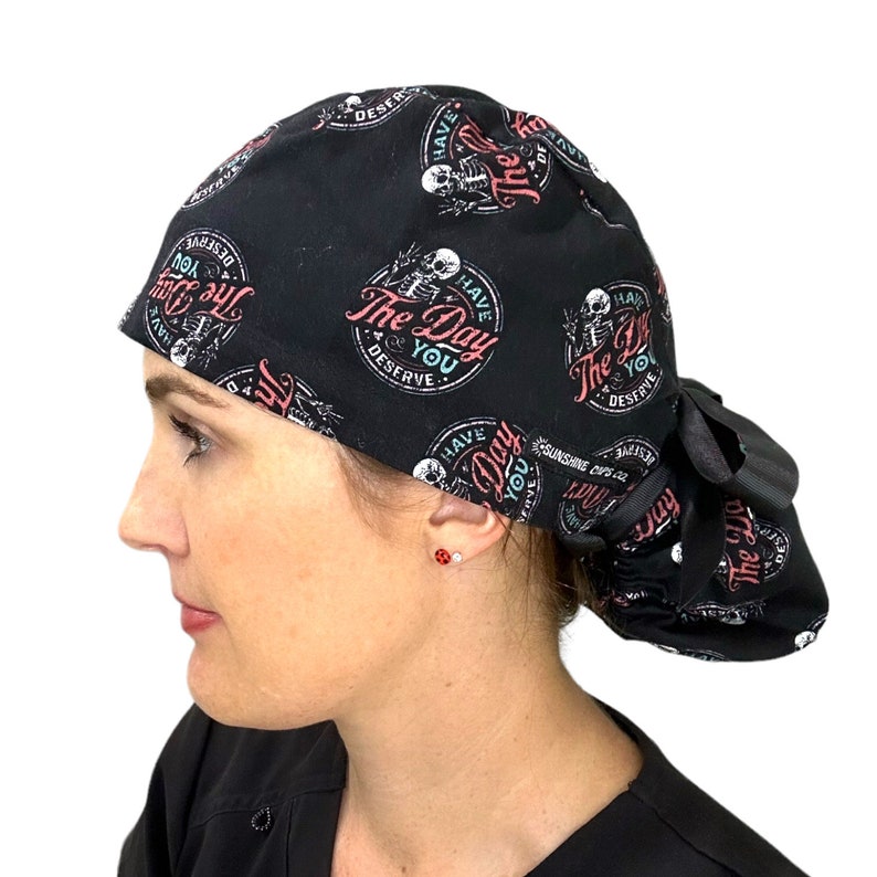 Puede incluir: Gorro quir&uacute;rgico negro con un patr&oacute;n repetido de un esqueleto y el texto "The Day You Deserve" en rosa y blanco. El gorro tiene la parte trasera fruncida y una cinta negra.