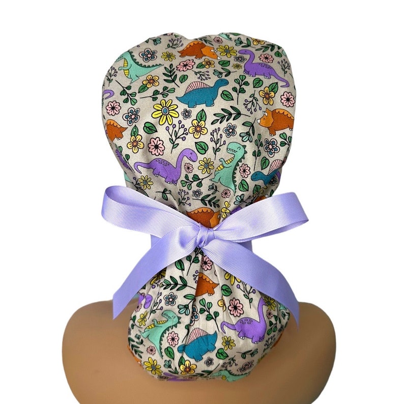 May include: A light purple scrub hat with a dinosaur and floral print. The hat has a tie that is tied in a bow at the back of the neck.