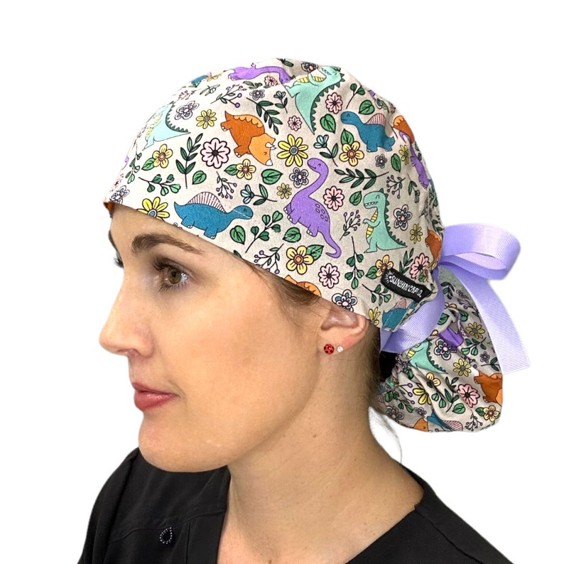May include: A light purple and white scrub hat with a dinosaur and floral print. The hat has a purple tie.