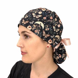 May include: A black surgical scrub cap with a floral and butterfly pattern. The cap features a crescent moon design and a light pink ribbon. The cap is designed to be worn during medical procedures.