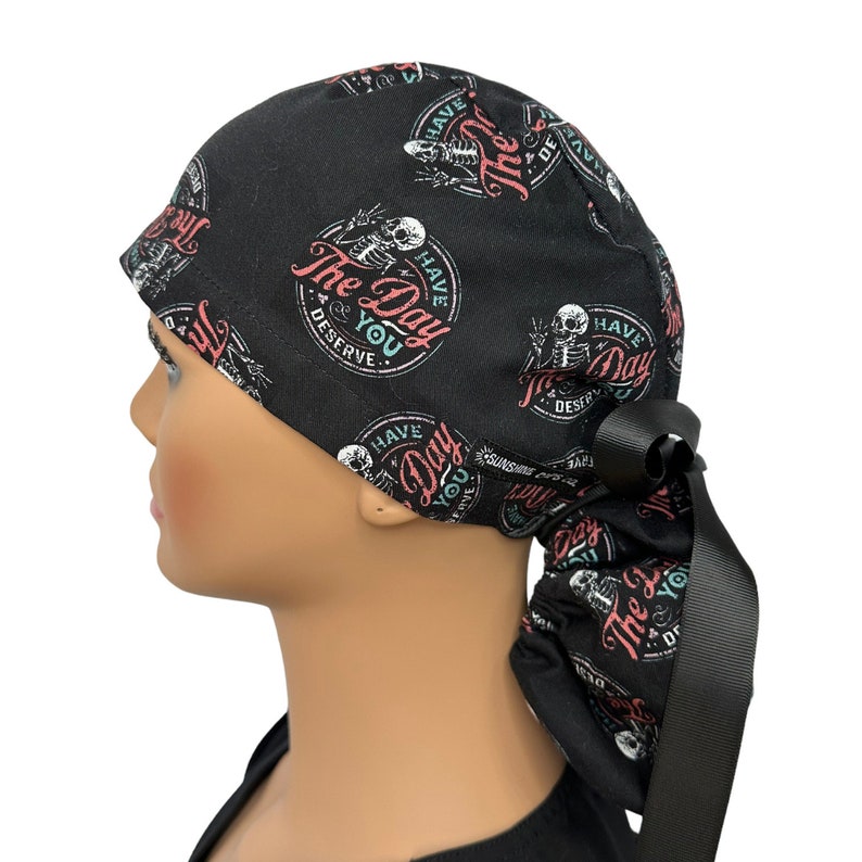 Puede incluir: Gorro quir&uacute;rgico negro con un patr&oacute;n repetido de esqueletos y el texto "Have The Day You Deserve" en un dise&ntilde;o circular. El gorro tiene una cinta negra. El dise&ntilde;o incluye acentos rosas, blancos y turquesas.