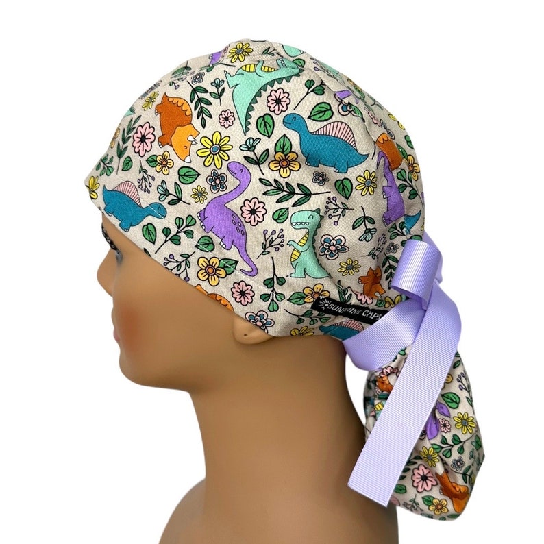 May include: A light beige scrub cap with a colorful dinosaur and floral print. The cap has a long lavender tie that is tied in a bow at the back.