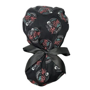 Puede incluir: Gorro de exfoliaci&oacute;n negro con un patr&oacute;n repetido de un esqueleto y el texto "Have The Day You Deserve" en rojo, verde azulado y blanco. El gorro est&aacute; atado con un lazo negro.