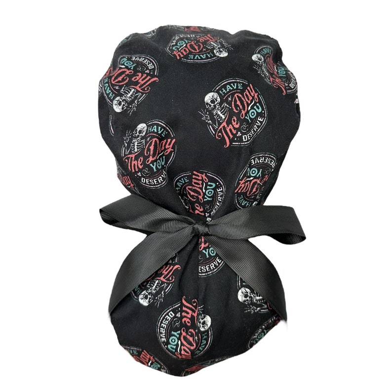 Puede incluir: Gorro de exfoliaci&oacute;n negro con un patr&oacute;n repetido de un esqueleto y el texto "Have The Day You Deserve" en rojo, verde azulado y blanco. El gorro est&aacute; atado con un lazo negro.