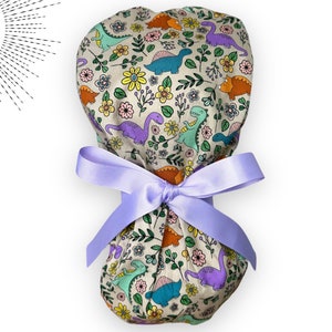 May include: A beige scrub hat with a colorful dinosaur and floral print. The hat is tied with a lavender ribbon.