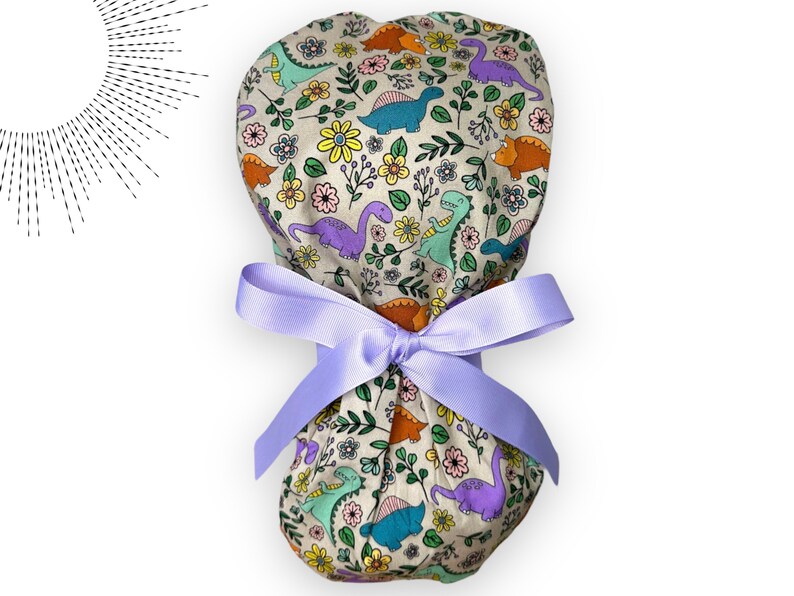 May include: A beige scrub hat with a colorful dinosaur and floral print. The hat is tied with a lavender ribbon.