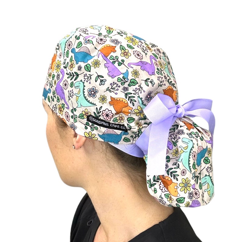 May include: A light purple scrub cap with a white background featuring a colorful dinosaur and floral print. The cap has a purple ribbon tied at the back. The text "Sunshine Caps Co." is printed on the cap.