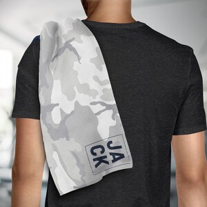 May include: A white and gray camouflage pattern towel with the name "JACK" printed in a navy blue font. The towel is draped over a person's shoulder, with a black t-shirt in the background. The towel is likely for use at the gym or during workouts.