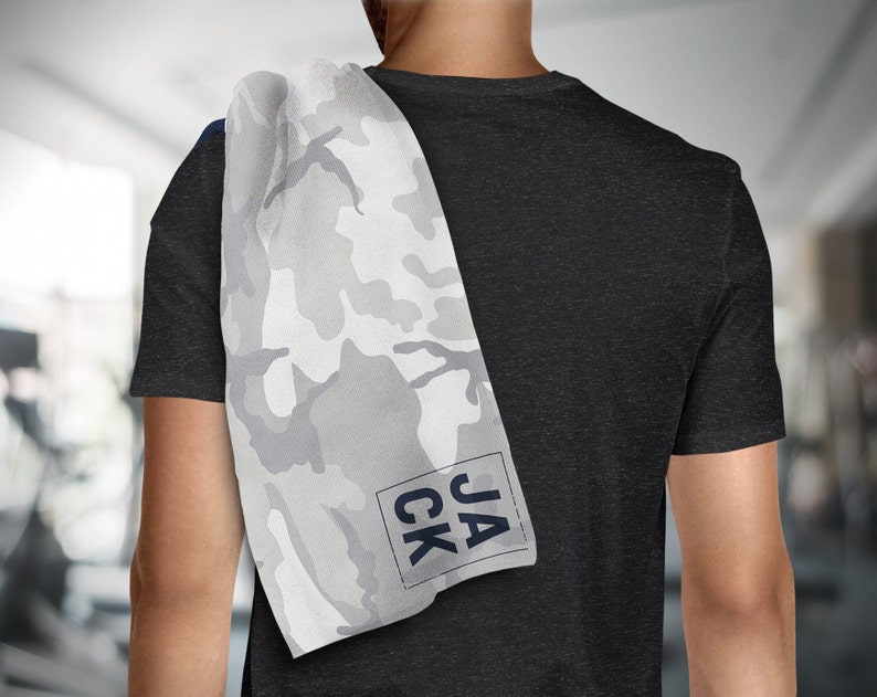 May include: A white and gray camouflage pattern towel with the name "JACK" printed in a navy blue font. The towel is draped over a person's shoulder, with a black t-shirt in the background. The towel is likely for use at the gym or during workouts.
