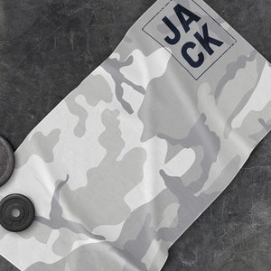 May include: A gray and white camouflage pattern workout towel with the name "JACK" printed in navy blue. Two black weight plates are visible in the image. The towel is folded and placed on a dark gray surface.