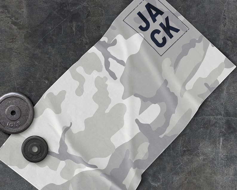 May include: A gray and white camouflage pattern workout towel with the name "JACK" printed in navy blue. Two black weight plates are visible in the image. The towel is folded and placed on a dark gray surface.