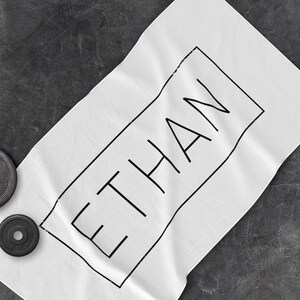 K&ouml;nnte beinhalten: Wei&szlig;es Handtuch mit dem Namen "ETHAN" in Schwarz in einem schwarzen Rechteck.