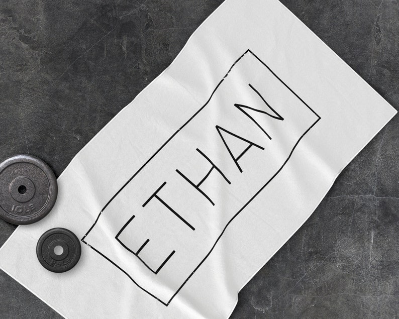 K&ouml;nnte beinhalten: Wei&szlig;es Handtuch mit dem Namen "ETHAN" in Schwarz in einem schwarzen Rechteck.