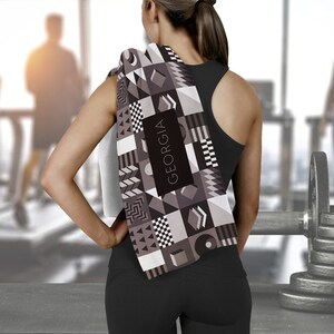 May include: A black and white geometric patterned towel with the word "GEORGIA" printed on it. The towel is draped over the shoulder of a person wearing a black tank top.
