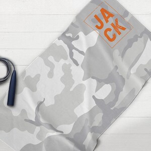 May include: A gray and white camouflage pattern towel with the name "JACK" in orange block letters. A gray jump rope with navy blue handles is next to the towel. The towel is folded on a white wooden surface.
