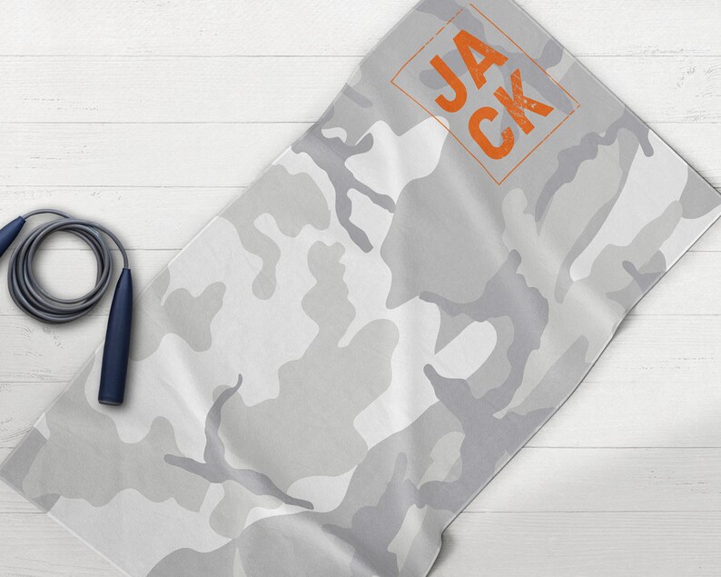 May include: A gray and white camouflage pattern towel with the name "JACK" in orange block letters. A gray jump rope with navy blue handles is next to the towel. The towel is folded on a white wooden surface.