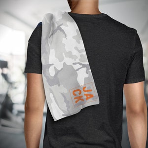 May include: A white and gray camouflage pattern towel with the name "JACK" in orange block letters. The towel is draped over a person's shoulder, with a dark gray t-shirt visible.