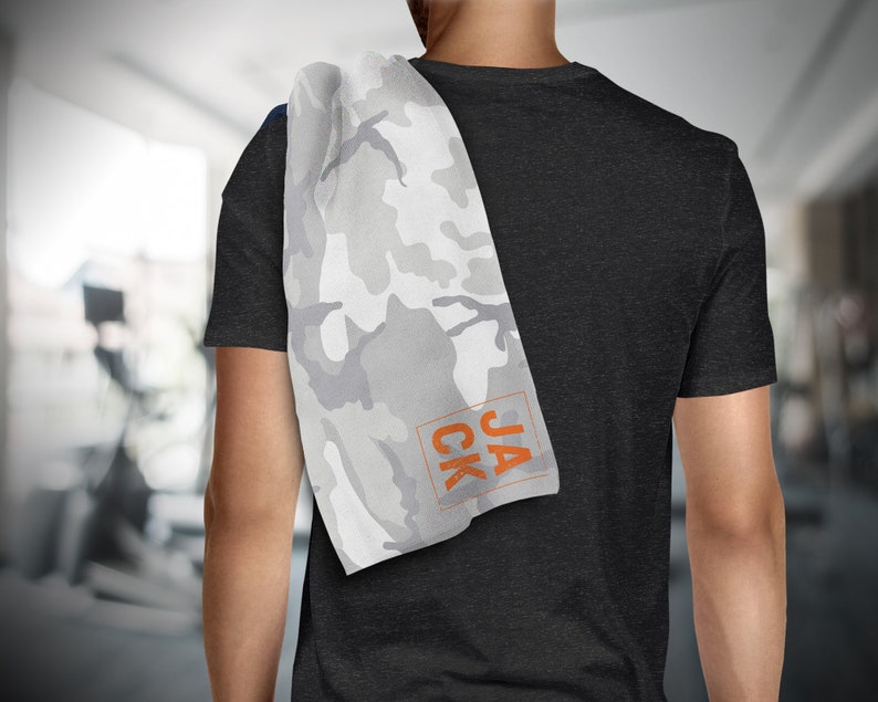 May include: A white and gray camouflage pattern towel with the name "JACK" in orange block letters. The towel is draped over a person's shoulder, with a dark gray t-shirt visible.