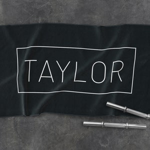 K&ouml;nnte beinhalten: Schwarzes Baumwollhandtuch mit dem Namen "TAYLOR" in wei&szlig; in einem wei&szlig;en Rechteck gedruckt.