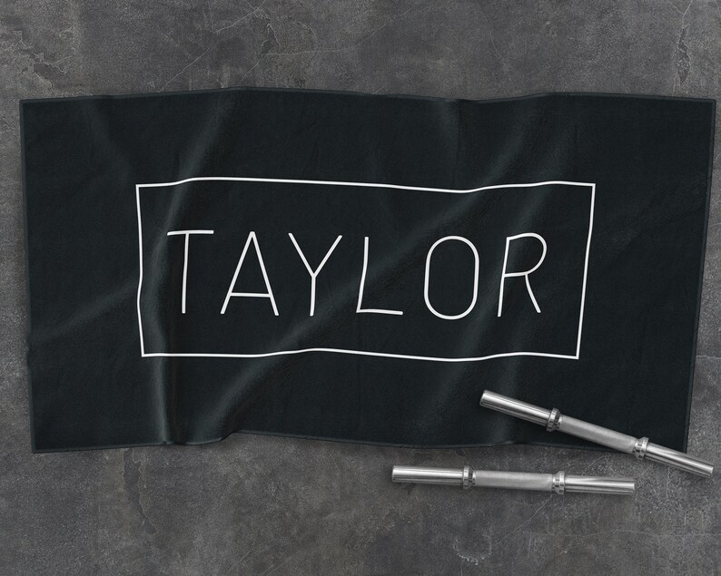 K&ouml;nnte beinhalten: Schwarzes Baumwollhandtuch mit dem Namen "TAYLOR" in wei&szlig; in einem wei&szlig;en Rechteck gedruckt.