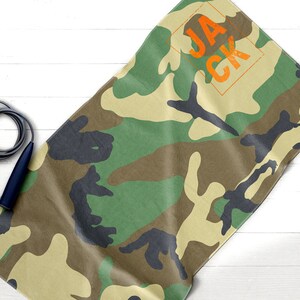 May include: Camouflage-patterned towel with the name "JACK" in orange block letters. A dark blue and gray jump rope is in the image. The towel has a green, brown, and black pattern.