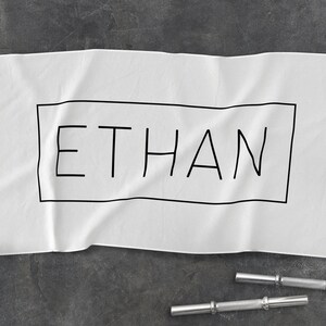 K&ouml;nnte beinhalten: Wei&szlig;es Baumwollhandtuch mit schwarzem Text, der "ETHAN" innerhalb eines schwarzen Rechtecks lautet.
