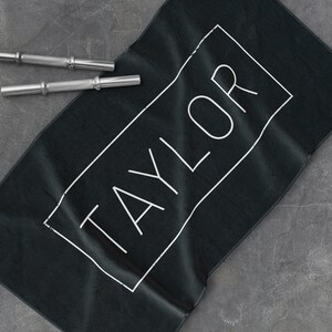 K&ouml;nnte beinhalten: Schwarzes Yogatuch mit dem Namen "TAYLOR" in wei&szlig; in einem wei&szlig;en Rechteck gedruckt.