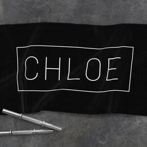 K&ouml;nnte beinhalten: Schwarzes Handtuch mit wei&szlig;em Rand und dem Wort "CHLOE" in wei&szlig; innerhalb des Randes.