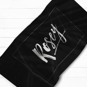 May include: Black beach towel with a white border and the name "Rosey" written in a silver script font.