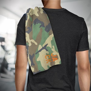 May include: A camouflage-patterned towel with green, brown, and black hues is draped over a person's shoulder. The towel has the orange text "JACK" in the corner. The background is a blurred gym setting.