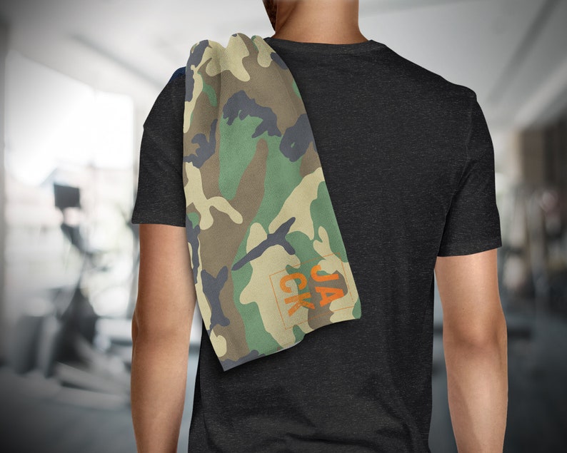 May include: A camouflage-patterned towel with green, brown, and black hues is draped over a person's shoulder. The towel has the orange text "JACK" in the corner. The background is a blurred gym setting.