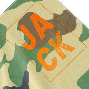 May include: Camouflage pattern fabric with the name "JACK" in orange block letters. The letters are inside an orange diamond shape. The fabric has green, brown, and tan colors.