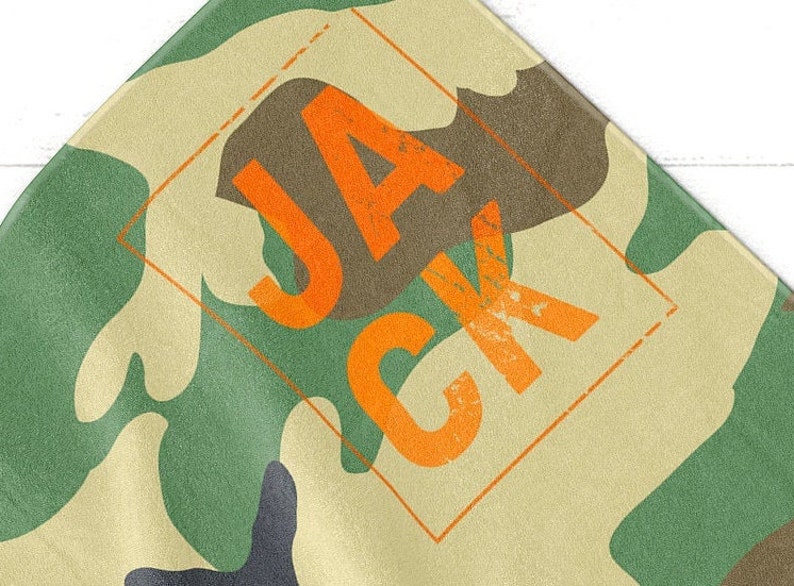 May include: Camouflage pattern fabric with the name "JACK" in orange block letters. The letters are inside an orange diamond shape. The fabric has green, brown, and tan colors.
