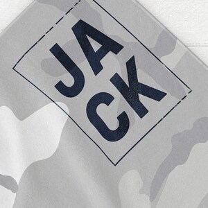 May include: A gray and white camouflage pattern textile with the word "JACK" in dark blue letters inside a dark blue rectangular frame. The fabric has a subtle, textured appearance.