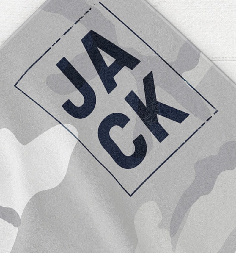 May include: A gray and white camouflage pattern textile with the word "JACK" in dark blue letters inside a dark blue rectangular frame. The fabric has a subtle, textured appearance.