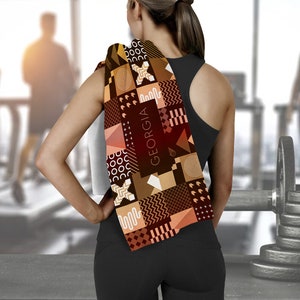 May include: A brown and tan patterned towel with the word "GEORGIA" printed on it. The towel is draped over the shoulder of a person wearing a black tank top and black pants.