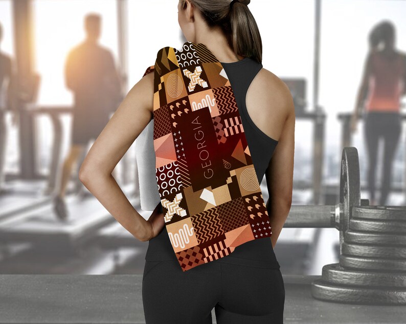 May include: A brown and tan patterned towel with the word "GEORGIA" printed on it. The towel is draped over the shoulder of a person wearing a black tank top and black pants.