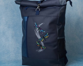 Könnte beinhalten: Marineblauer Rucksack mit Rollverschluss und gesticktem Libellen-Design. Der Rucksack hat eine Fronttasche mit Reißverschluss, eine seitliche Netztasche und einen silberfarbenen Verschluss. Die Libellen haben farbige Flügel.