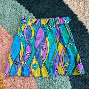 May include: A mini skirt featuring a vibrant, multi-colored wavy pattern in shades of blue, green, purple, and yellow. The skirt is adorned with small white and blue flower motifs and includes a matching fabric belt with a rectangular buckle.