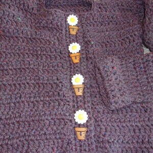 May include: A purple crocheted cardigan with five white daisy buttons. Each button has a brown pot.