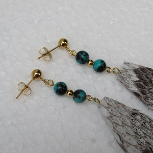 Op de afbeelding: Een paar oorhangers met turquoise en zwart gepatroneerde kralen en een grijs en wit slangenhuid hanger.